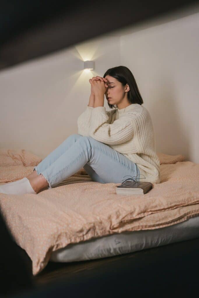Woman in White Sweater and Blue Denim Jeans Praying on Bed Photo used as a featured image for Are Novenas Superstitious in About Catholics Blog